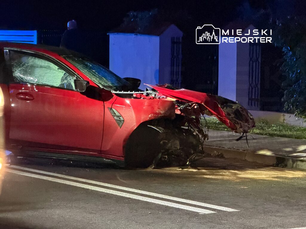 Czerwony samochód osobowy z uszkodzoną przednią częścią stoi na poboczu drogi nocą, w tle widać jasne światła.