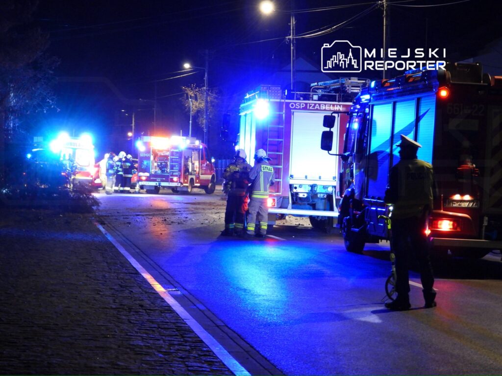 Grupa strażaków i policjantów pracuje przy wozach ratunkowych na oświetlonej nocą drodze.