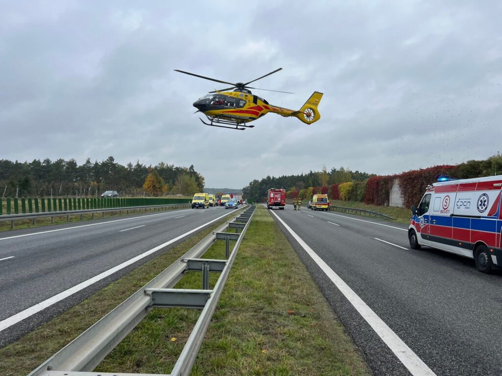 Śmigłowiec ratunkowy unosi się nad autostradą, obok zespoły ratunkowe i pojazdy służb medycznych.