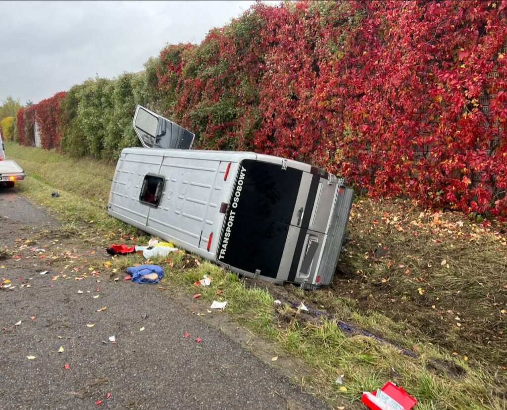 Przewoźnik osobowy leży na boku przy drodze, otoczony kolorowymi liśćmi i ubraniami na poboczu.