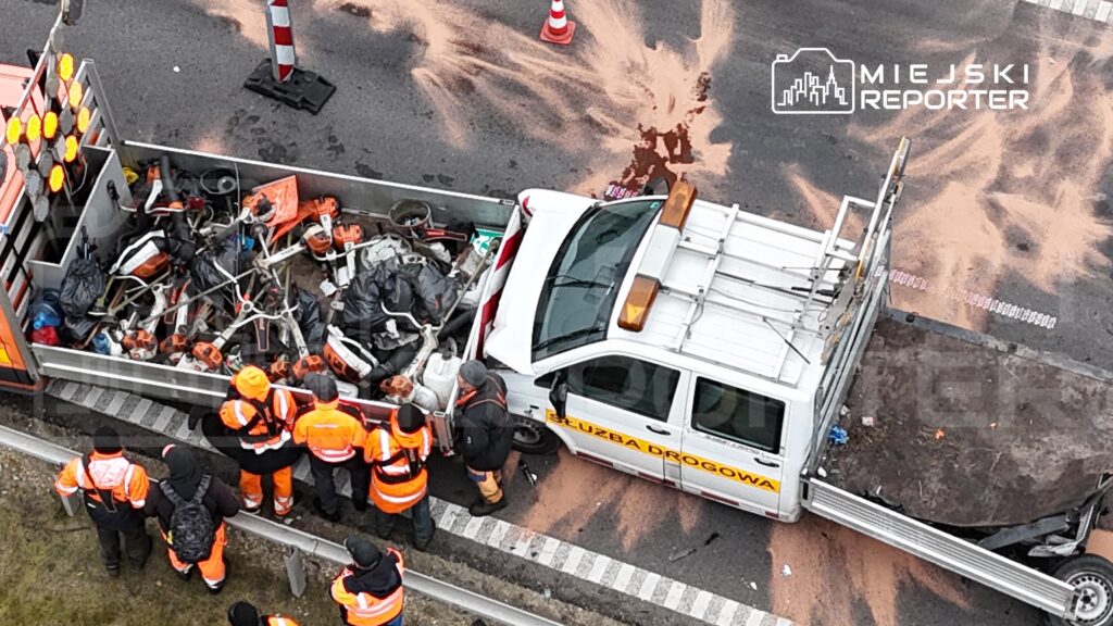 Grupa pracowników w pomarańczowych kurtkach zbiera rozbite elementy na poboczu drogi, obok policyjnego samochodu.