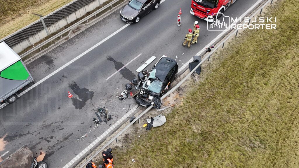 Na drodze leży uszkodzony samochód osobowy, wokół niego pracują strażacy w mundurach. Obok widać rozbite części pojazdu.
