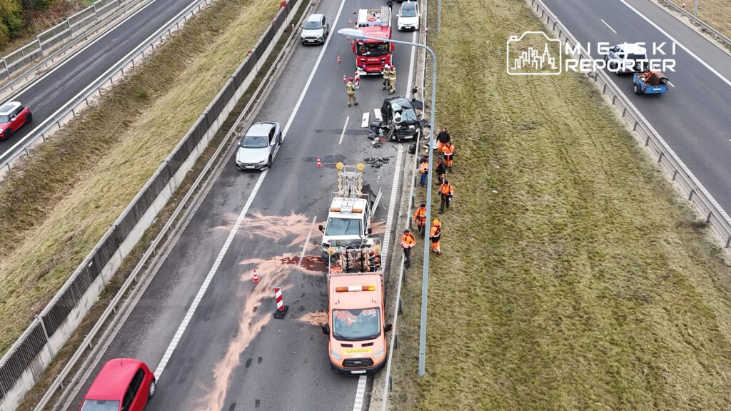 Na autostradzie pracownicy w pomarańczowych uniformach sprzątają miejsce wypadku, obok stoi uszkodzony samochód.