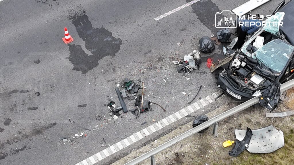 Wrak samochodu osobowego z rozbitą szybą leży na poboczu drogi, dookoła porozrzucane fragmenty pojazdu i przedmioty.