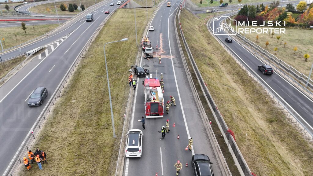 Grupa strażaków i ratowników pracuje przy wypadku drogowym na pasie ruchu, obok uszkodzonego pojazdu.