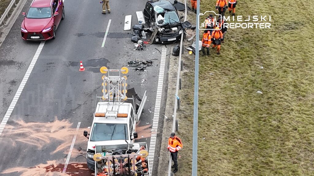 Na drodze leży rozbity samochód osobowy, a obok pracują służby ratunkowe w pomarańczowych ubraniach.