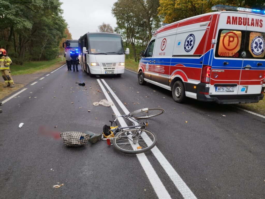 Na drodze leży przewrócony rower obok torby, a w tle widać ambulans oraz pojazd z niebieskimi światłami.