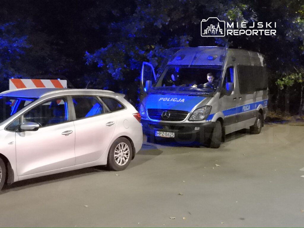 Policyjny radiowóz z niebieskimi światłami stoi obok zaparkowanego samochodu osobowego na nocnej ulicy.