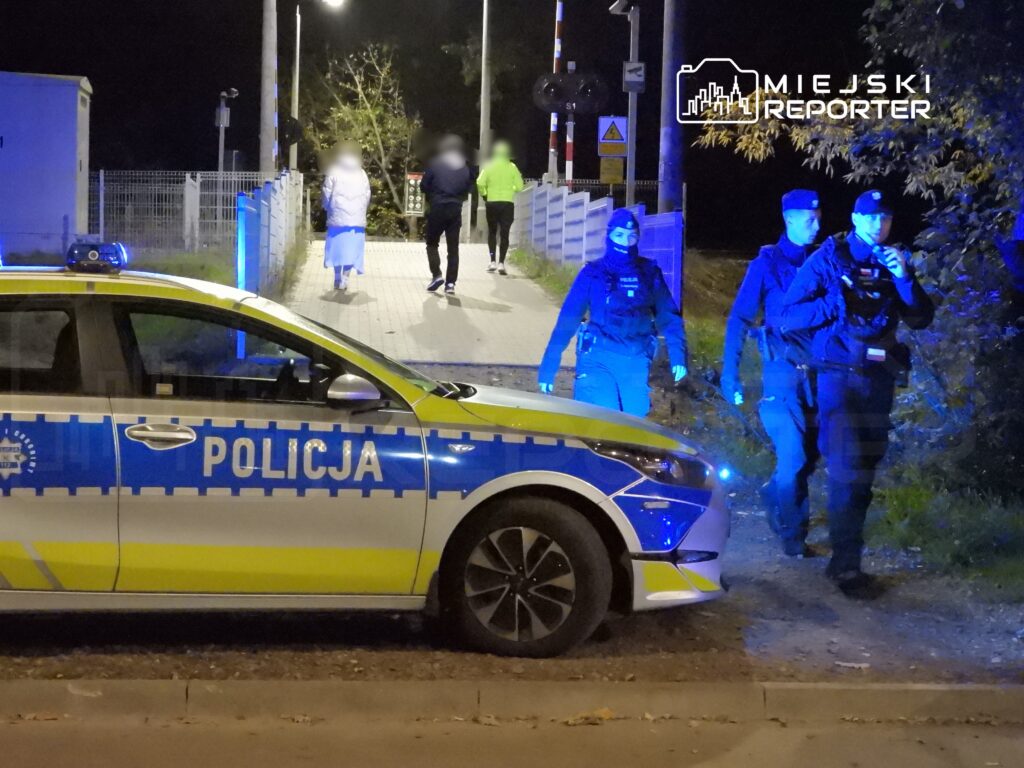 Policjanci w mundurach patrolują okolicę nocą, obok radiowozu z włączonymi światłami, w tle piesi przechodzą przez most.