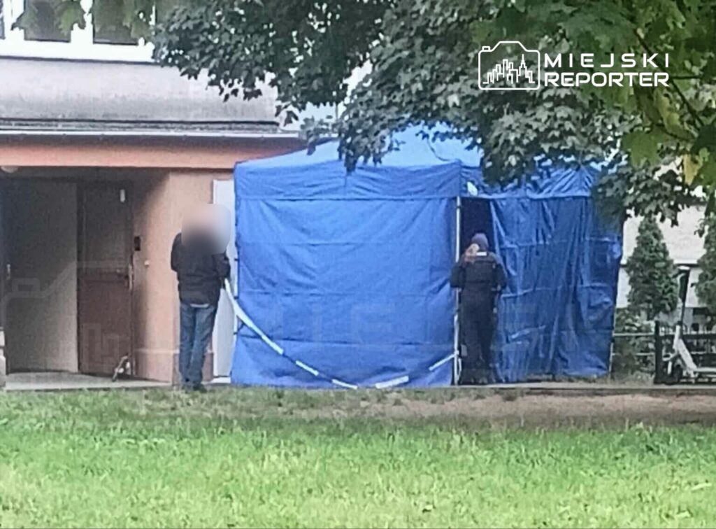Mężczyzna w czarnej kurtce stoi obok niebieskiego namiotu, a obok niego policjant w mundurze wchodzi do środka.