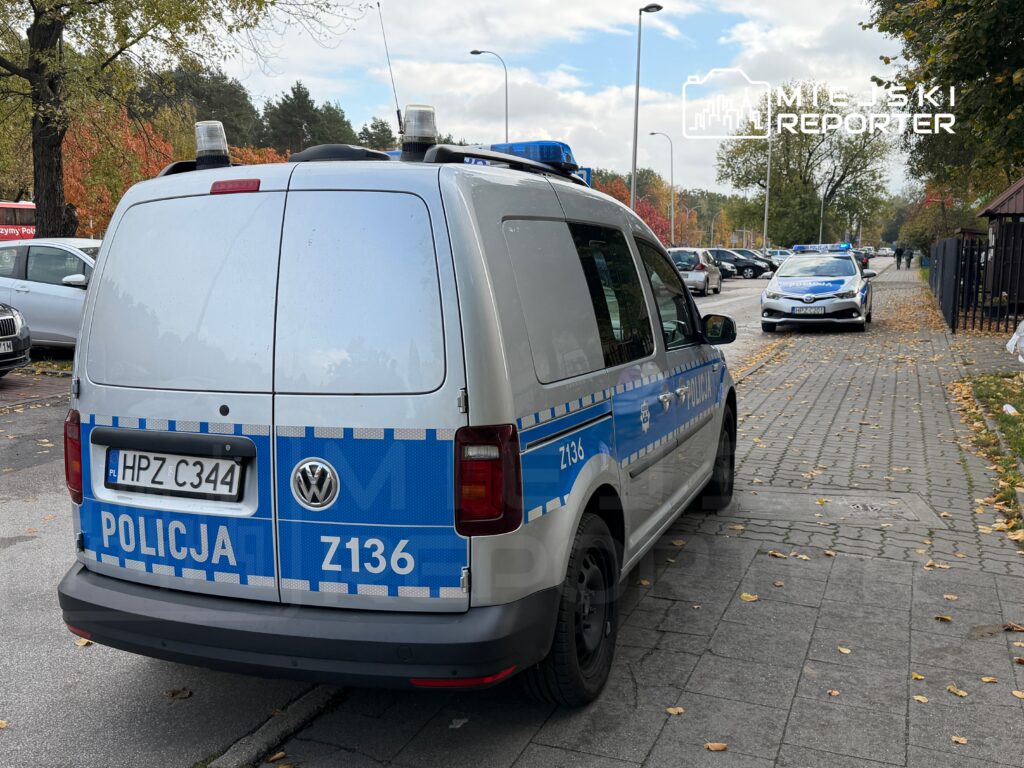 Radiowóz policyjny zaparkowany na chodniku, obok inny pojazd służbowy, w tle drzewa i zaparkowane samochody.