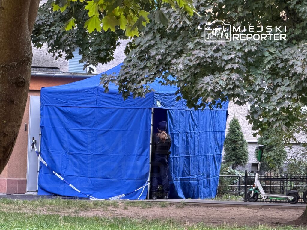 Funkcjonariusz policji w mundurze wchodzi do niebieskiego namiotu postawionego w parku.