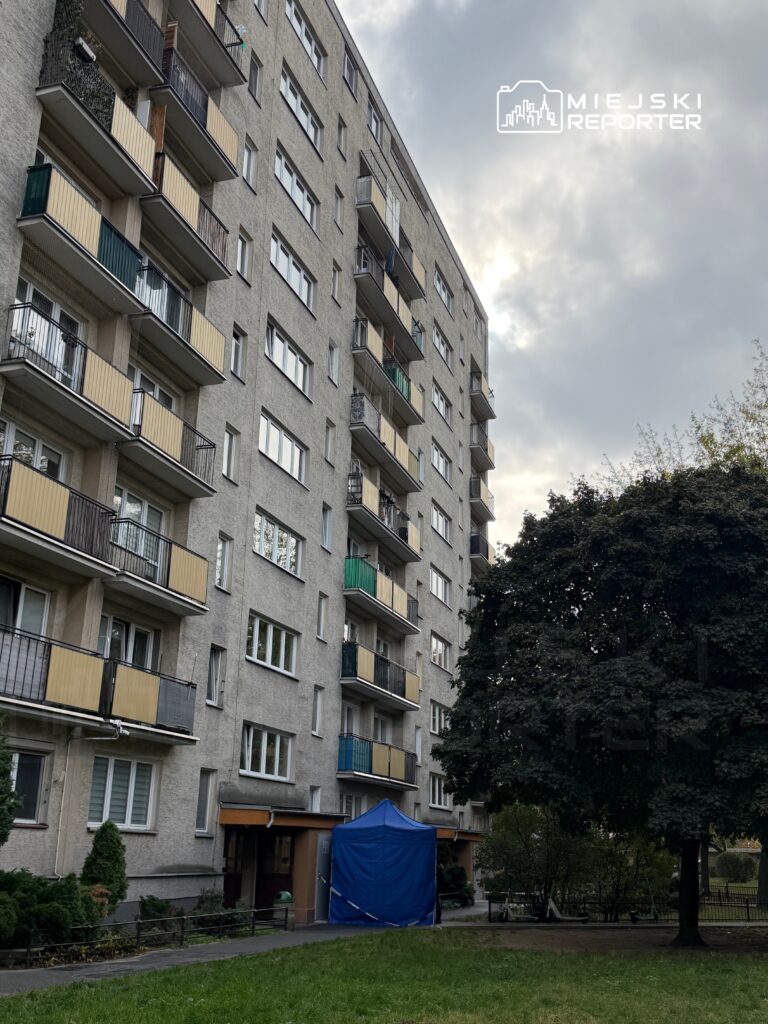 Siedmiopiętrowy blok mieszkalny z balkonami, z niebieskim namiotem przed wejściem, otoczony zielenią.