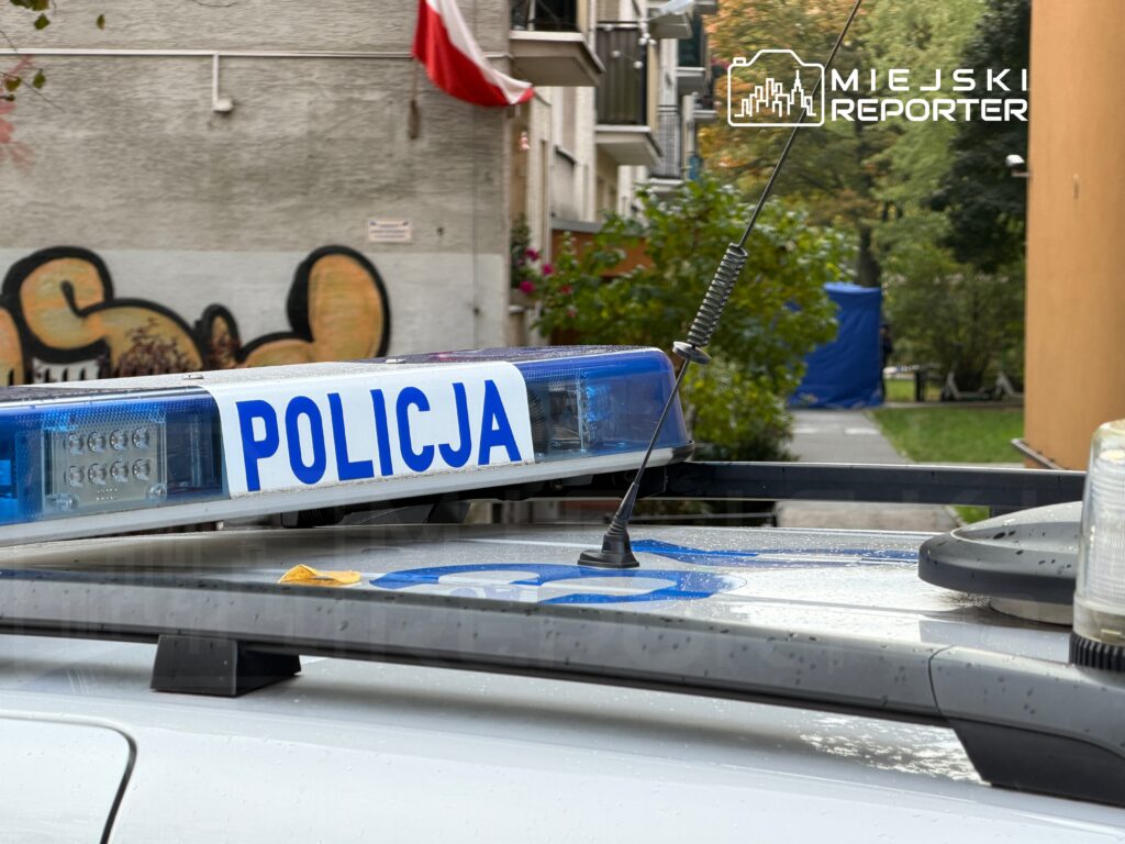 Policyjny radiowóz z niebieską sygnalizacją zaparkowany na ulicy, w tle widoczne graffiti i budynki mieszkalne.