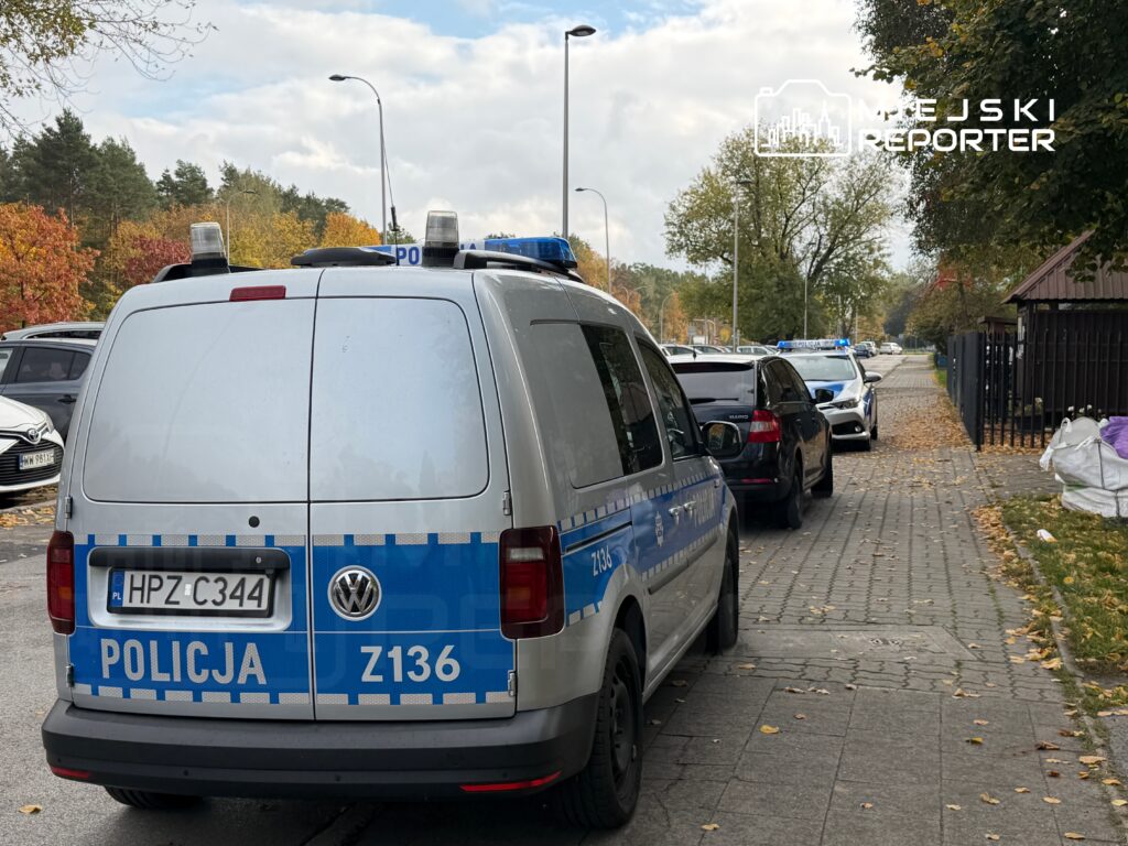 Policyjny samochód dostawczy zaparkowany na ulicy, obok stoją inne pojazdy, w tle widać drzewa z jesiennymi liśćmi.