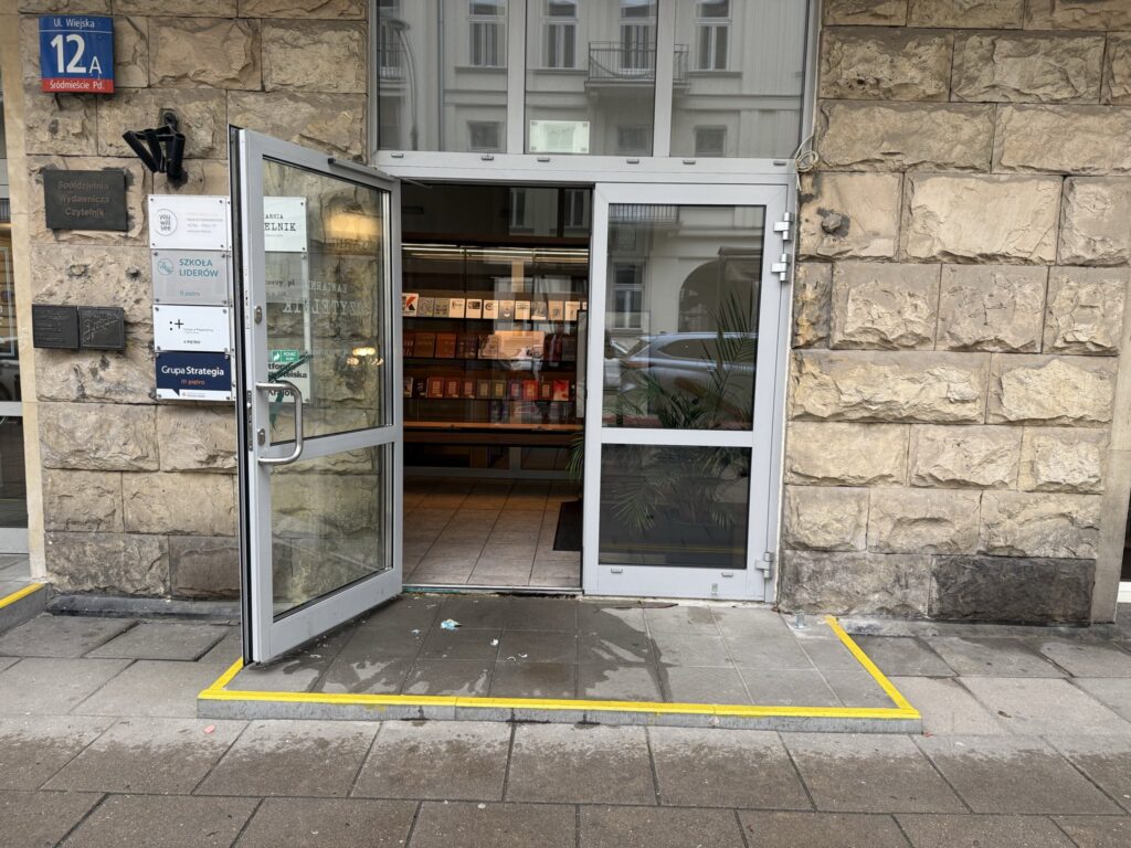 Drzwi wejściowe do budynku z kamienną elewacją, z widocznym znakiem "U. Wiejska 12A" oraz żółtą linią na chodniku.