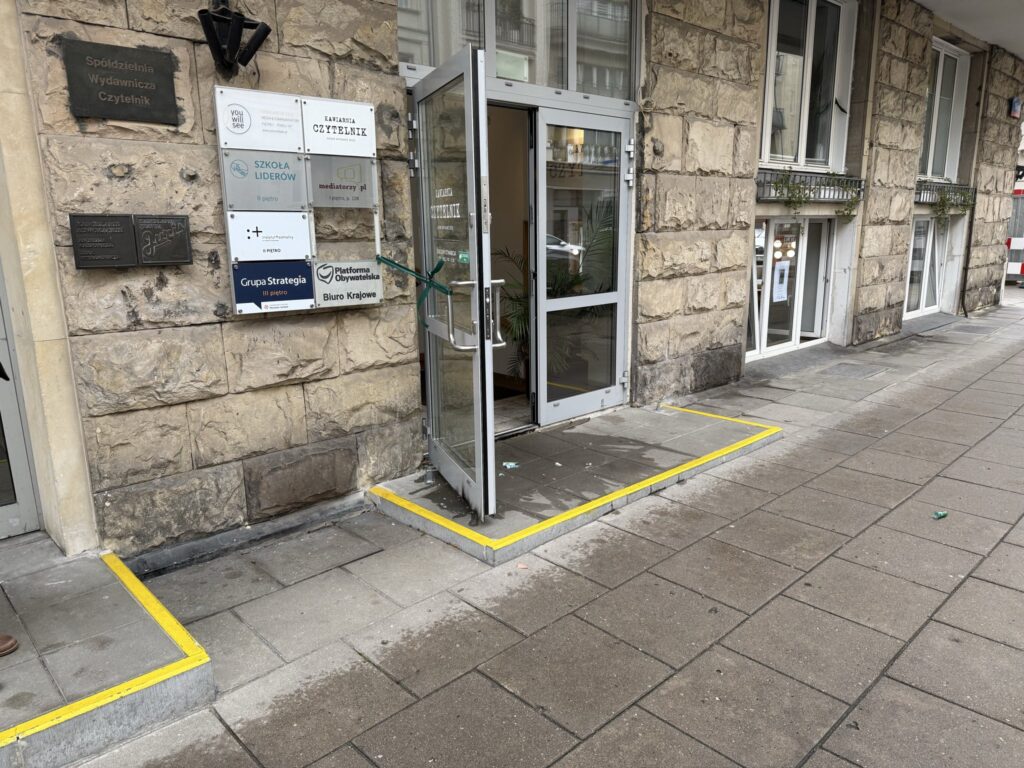 Drzwi wejściowe do budynku z kamienną elewacją, z oznaczeniem "Czytelnik" i innymi tabliczkami informacyjnymi.