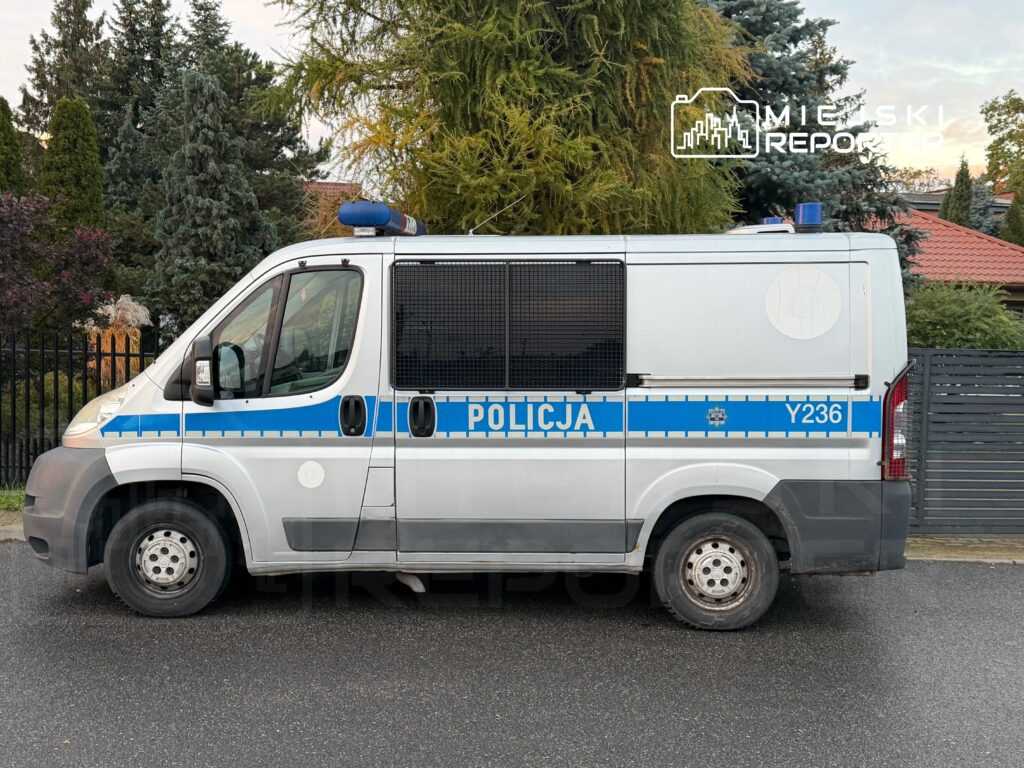 Funkcjonariusz policji w radiowozie parkowanym na ulicy, w tle ogrodzenie i drzewa.