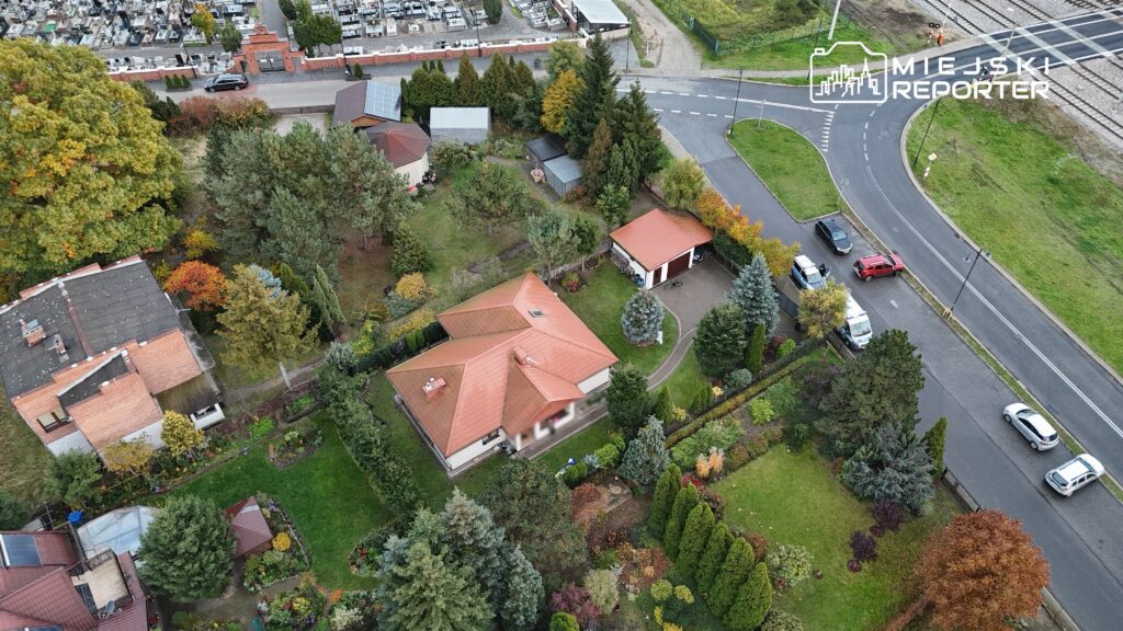 Widok z lotu ptaka na dom jednorodzinny z czerwonym dachem otoczony zielonym ogrodem i drzewami, w tle cmentarz.