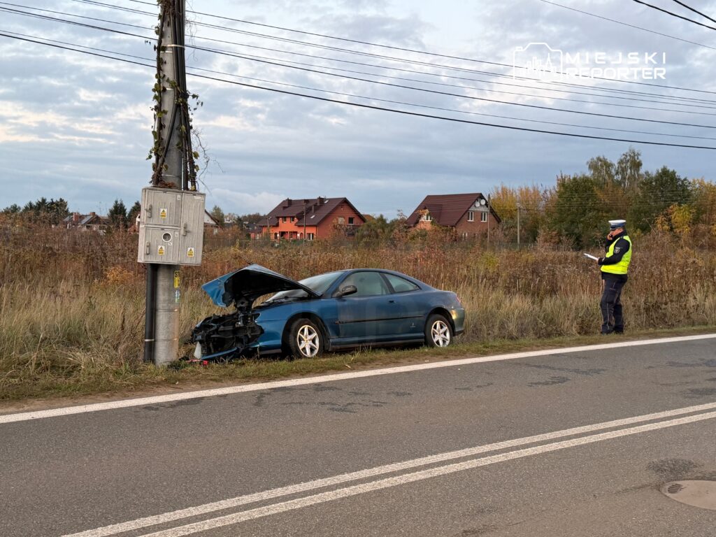 Nieoznakowany samochód osobowy z uszkodzoną maską stoi przy słupie energetycznym, obok policjant sporządzający notatki.