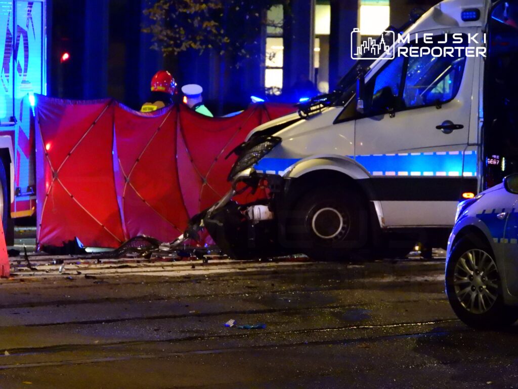 Wypadek drogowy z udziałem pojazdu dostawczego i barier ochronnych, w tle niebieskie światła służb ratunkowych.