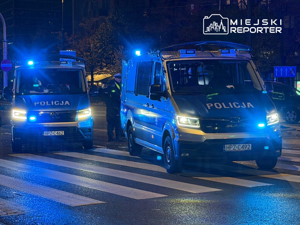 Policyjne samochody z niebieskimi światłami stoją na przejściu dla pieszych, a policjant rozmawia z innym funkcjonariuszem.