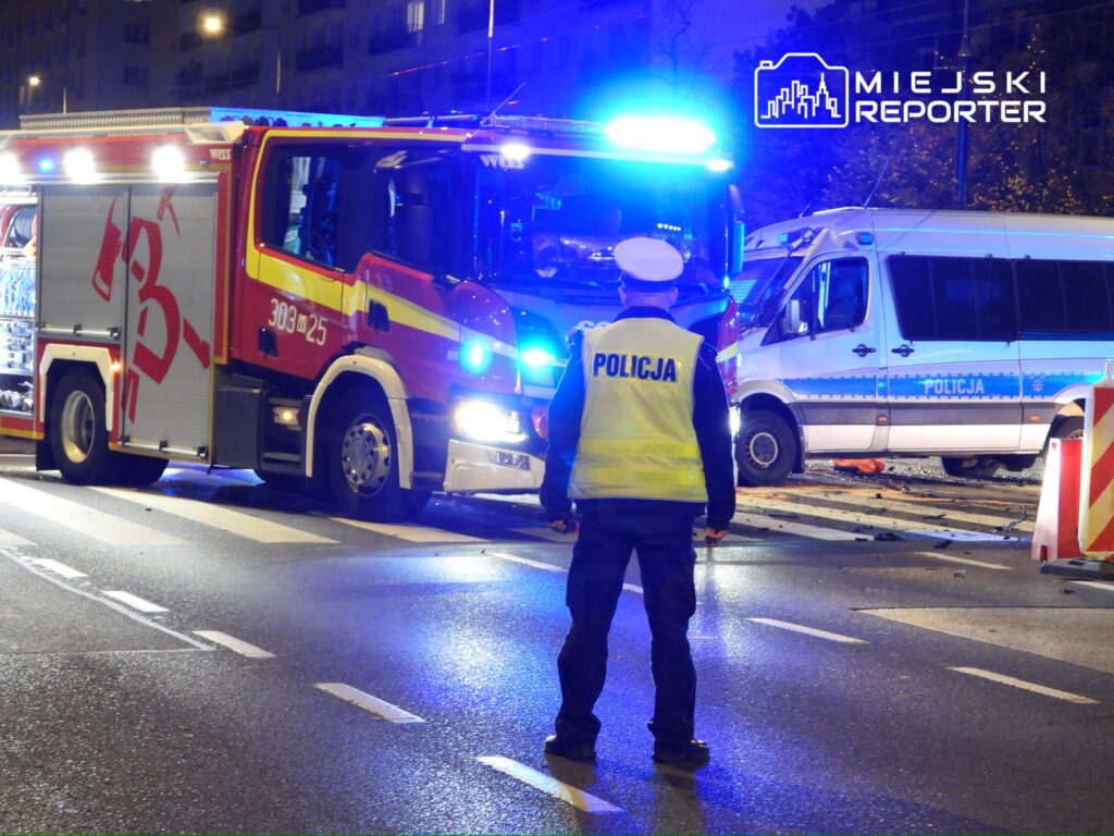 Strażak przy wozie strażackim z włączonymi sygnalizacjami, a obok policjant kierujący ruchem na oświetlonej ulicy.