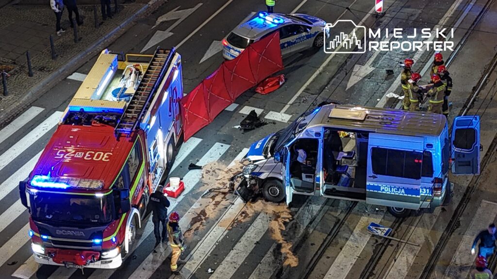 W nocy na ulicy zablokowanej przez czerwony parawan, strażacy i policjanci pracują przy uszkodzonym pojeździe.
