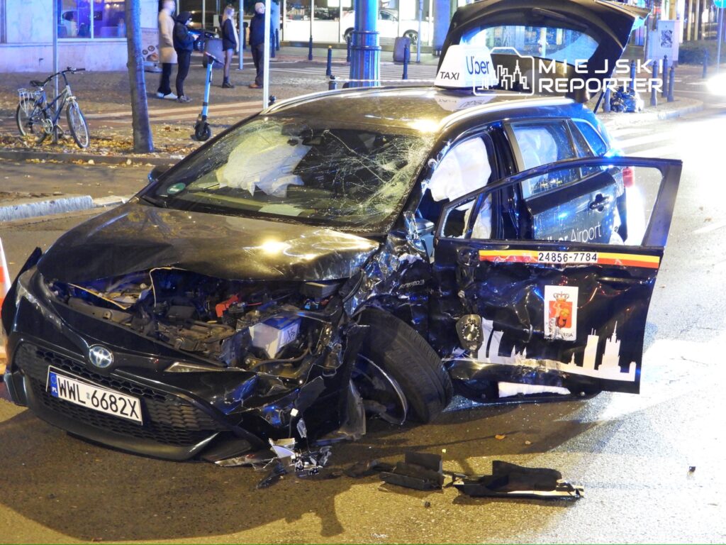 Uszkodzony samochód osobowy z otwartymi drzwiami stoi na ulicy, obok widać rowery i przechodniów.