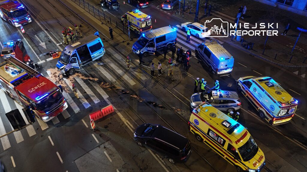 Na skrzyżowaniu ulicy stoją pojazdy służb ratunkowych, w tym ambulansy i radiowozy, a strażacy udzielają pomocy poszkodowanym.