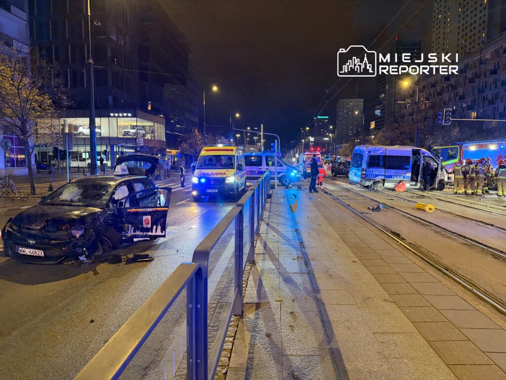 W nocy na ulicy widać uszkodzony samochód osobowy, obok niego policyjny radiowóz oraz ambulans, a w tle strażacy pomagają poszkodowanym.