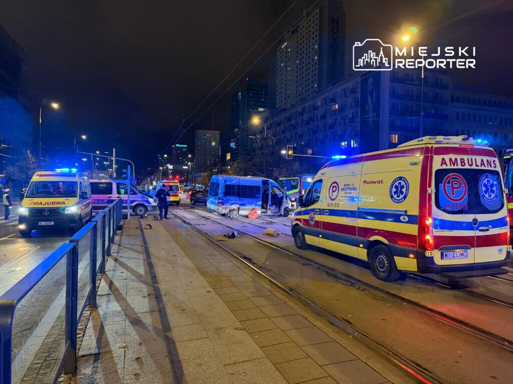 Na ulicy, w nocy, zespoły ratunkowe i policja interweniują po wypadku drogowym. W tle widać nowoczesne budynki.