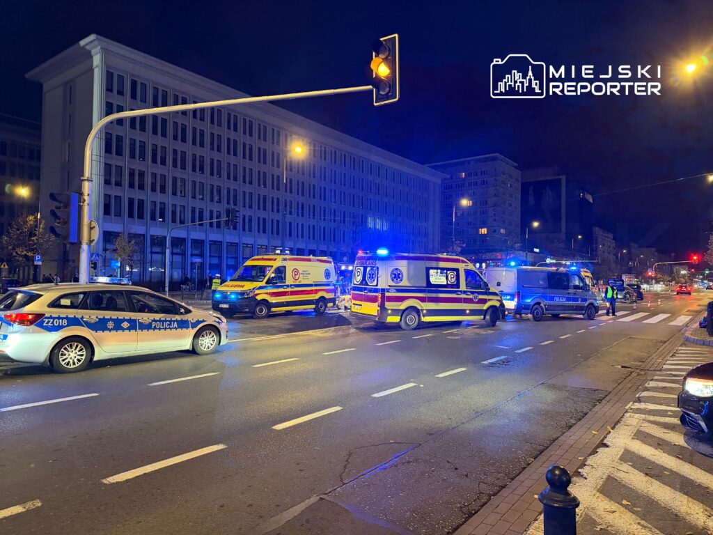 Policja i karetki pogotowia na oświetlonej ulicy, w tle nowoczesny biurowiec. Ruch wstrzymany.