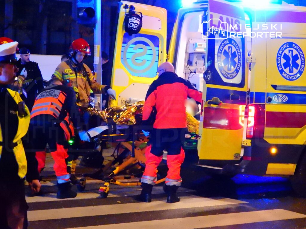 Ratownicy medyczni przenoszą poszkodowanego na noszach do karetki w nocy, w otoczeniu policyjnym.