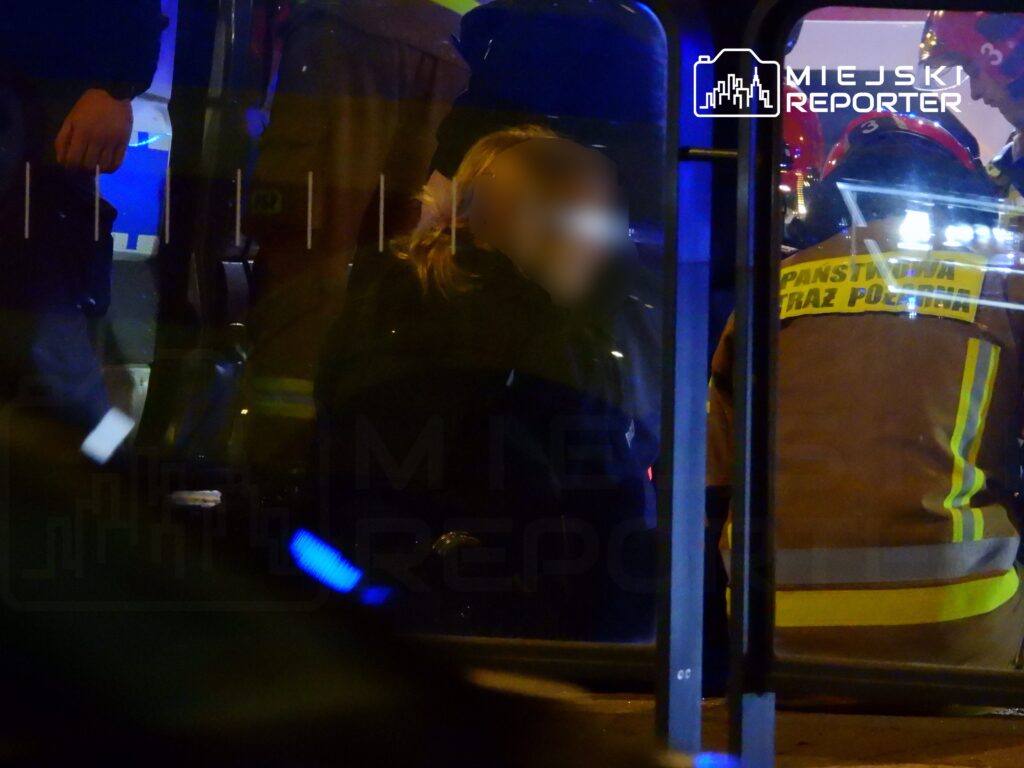 Kobieta w ciemnym ubraniu siedzi w pojeździe, podczas gdy strażacy w mundurach udzielają jej pomocy.