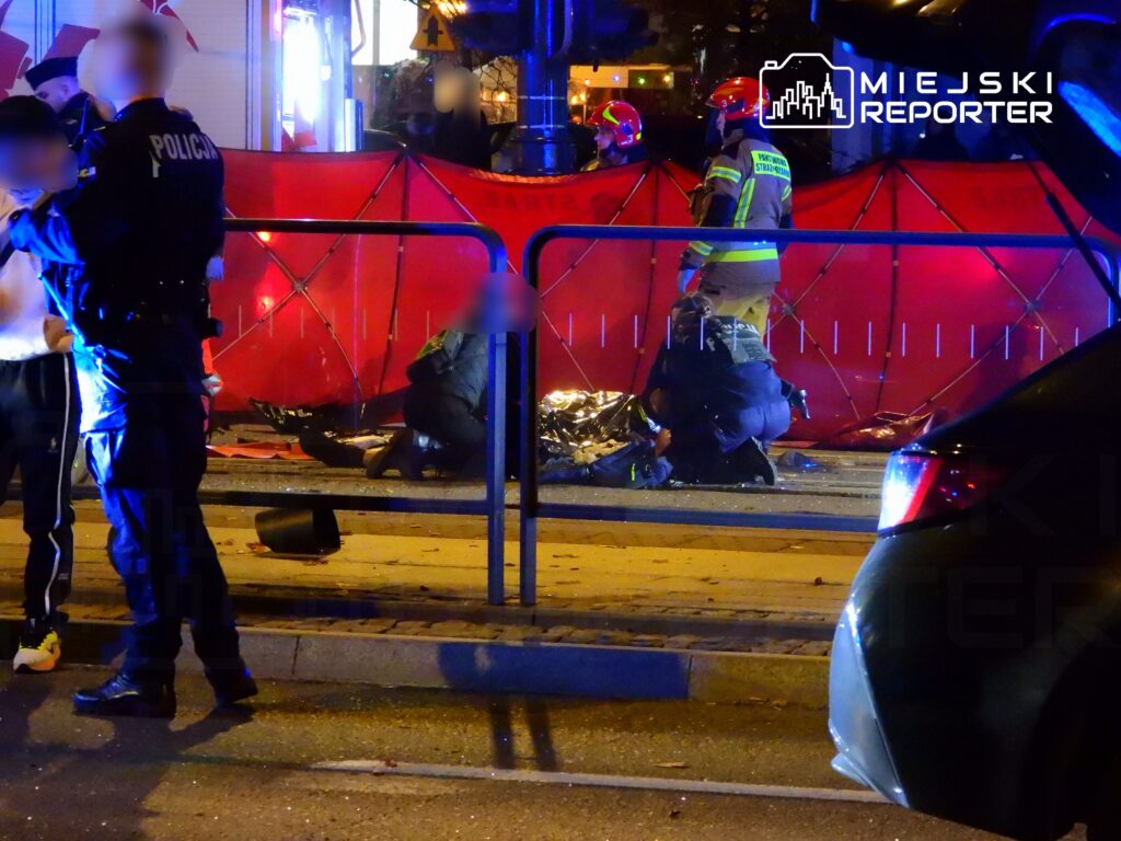 Policjanci i strażacy udzielają pomocy poszkodowanemu na ulicy, w tle widoczna czerwona osłona zabezpieczająca miejsce zdarzenia.