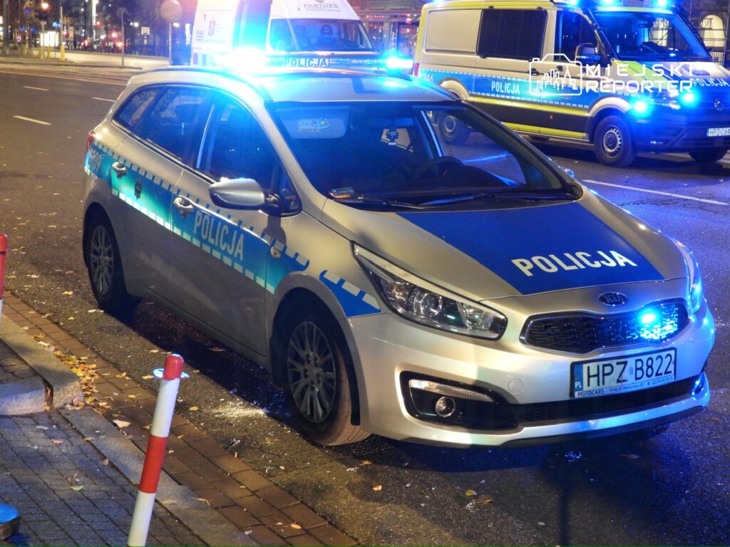 Radiowóz policyjny z włączonymi sygnałami świetlnymi stoi na ulicy nocą, obok drugiego pojazdu służb mundurowych.