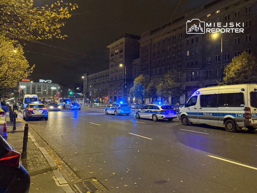 Policyjne samochody z włączonymi sygnałami świetlnymi zatrzymane na ruchliwej ulicy nocą, w tle oświetlony budynek.