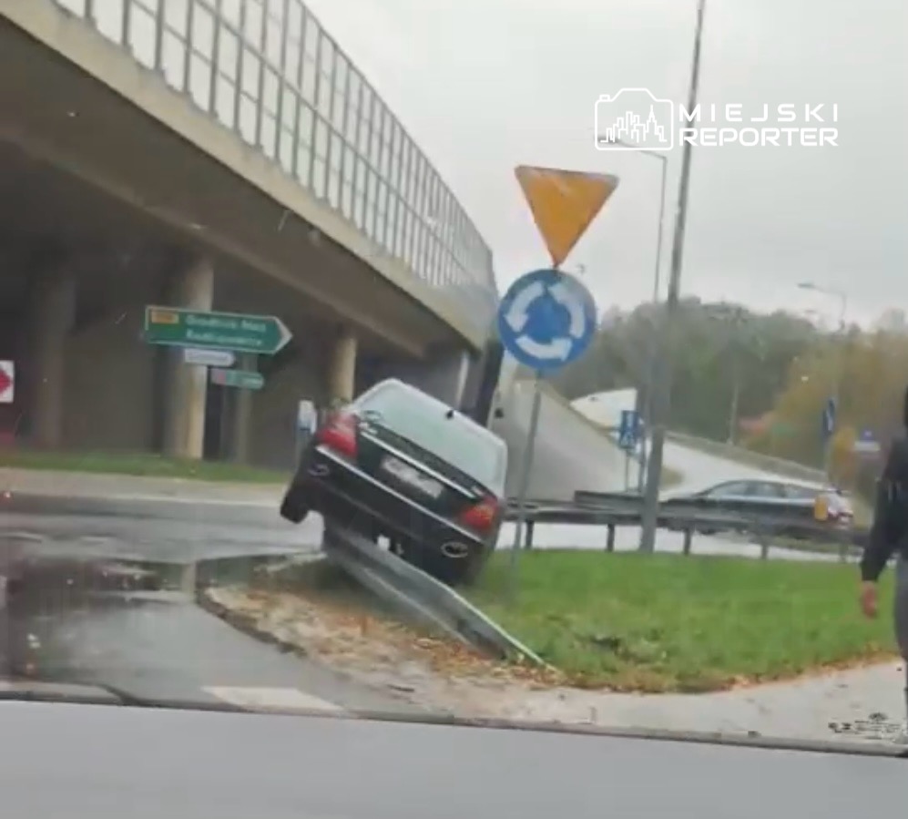 Czarny samochód osobowy unosi się na krawędzi drogi przy rondzie, obok znaku drogowym.