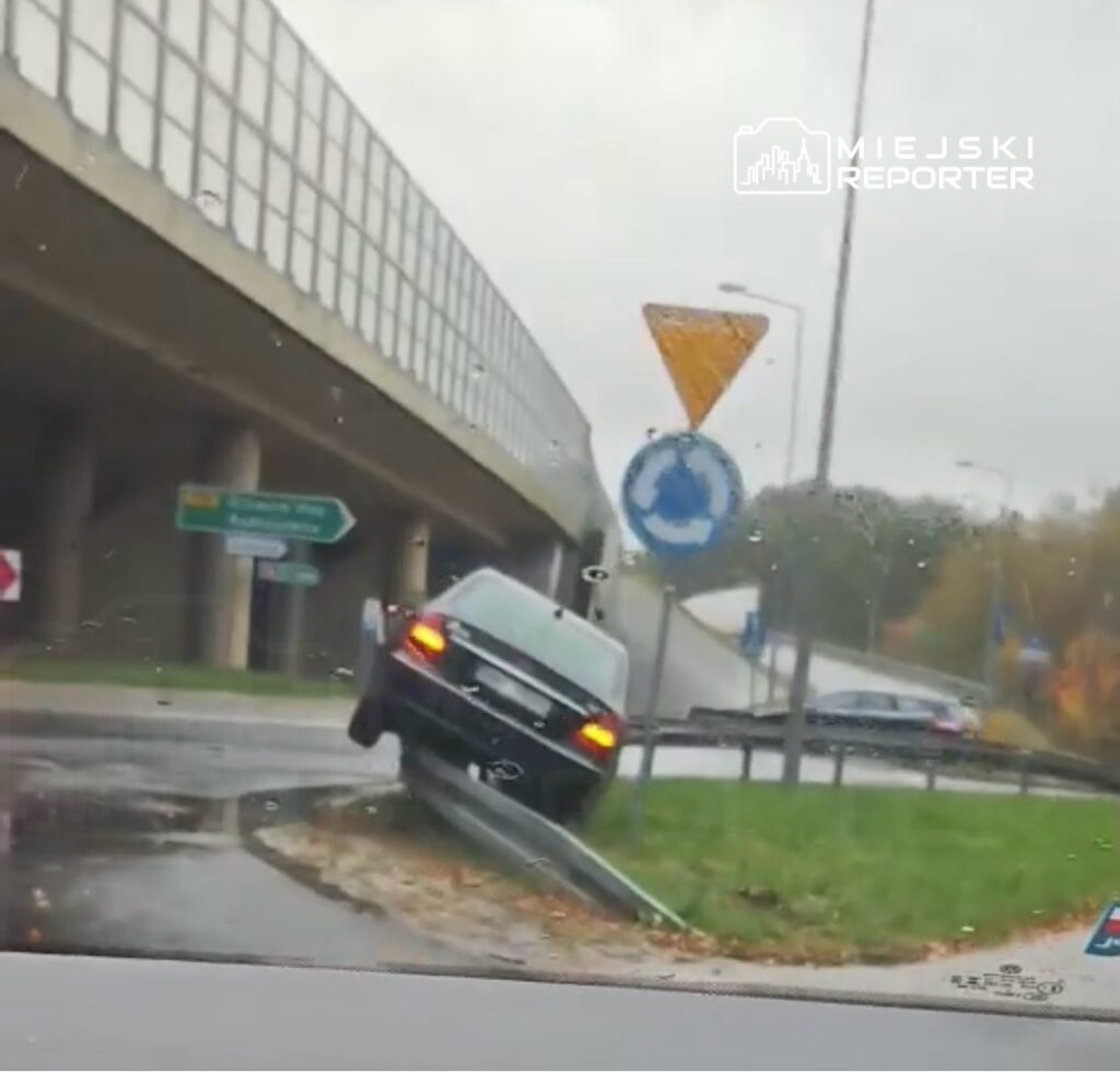 Czarny samochód osobowy unosi się na rondzie, z jednym kołem na krawężniku, w deszczowej aurze.