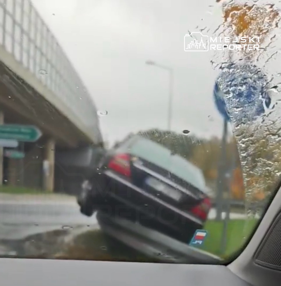 Samochód osobowy stoi na krawężniku, częściowo uniesiony, w deszczowej aurze, obok znaku drogowym.