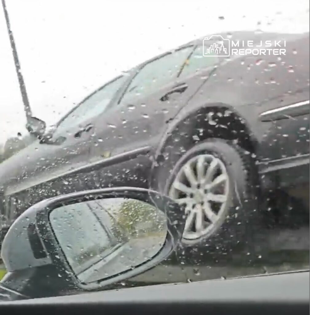Czarny samochód osobowy porusza się po drodze w deszczową pogodę, widoczny przez mokre szyby innego pojazdu.