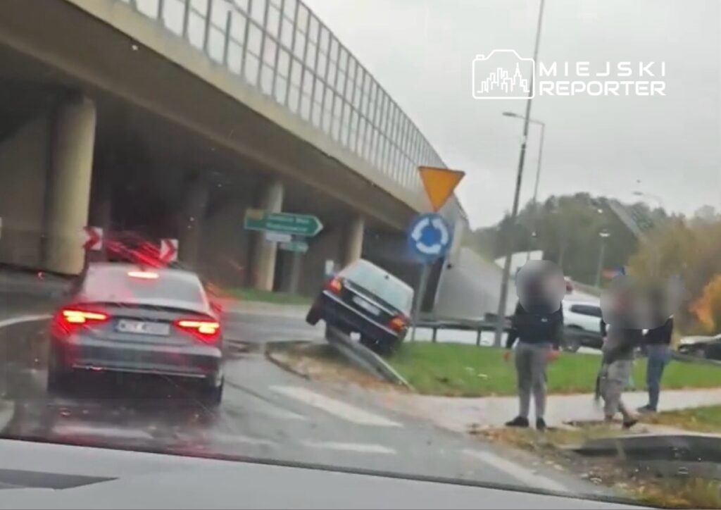 Czarny samochód unosi się na krawędzi ronda, obok trzech osób stojących na chodniku w deszczowej pogodzie.