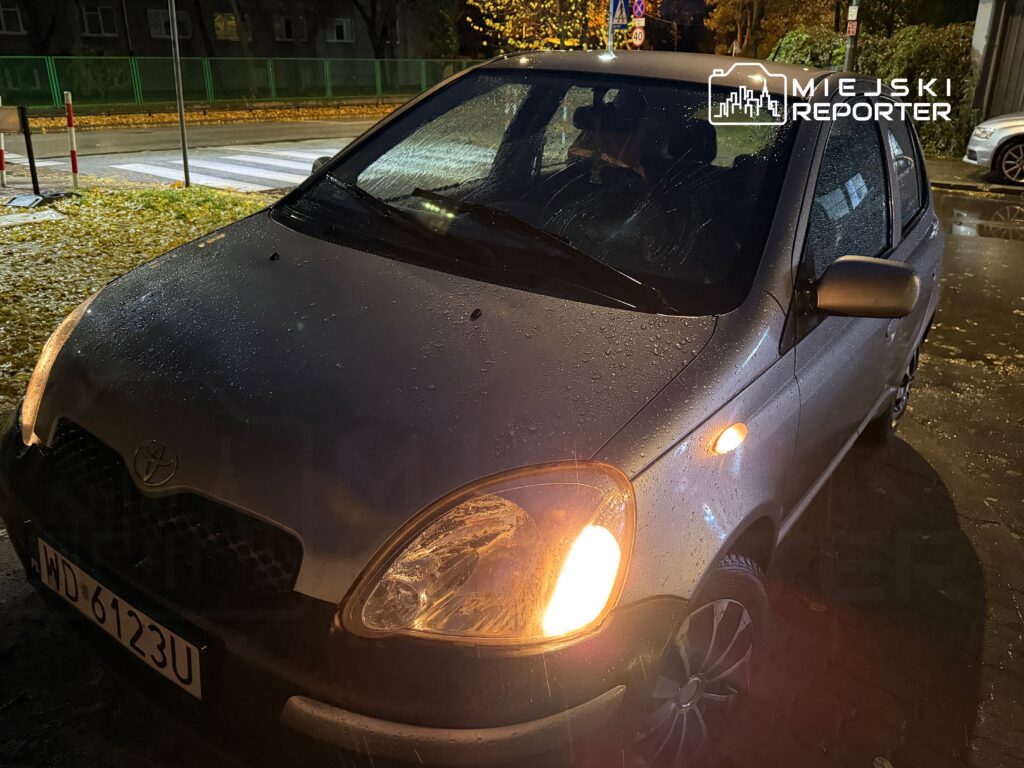 Samochód osobowy marki Toyota zaparkowany na mokrej ulicy, z kroplami wody na karoserii i włączonym światłem.