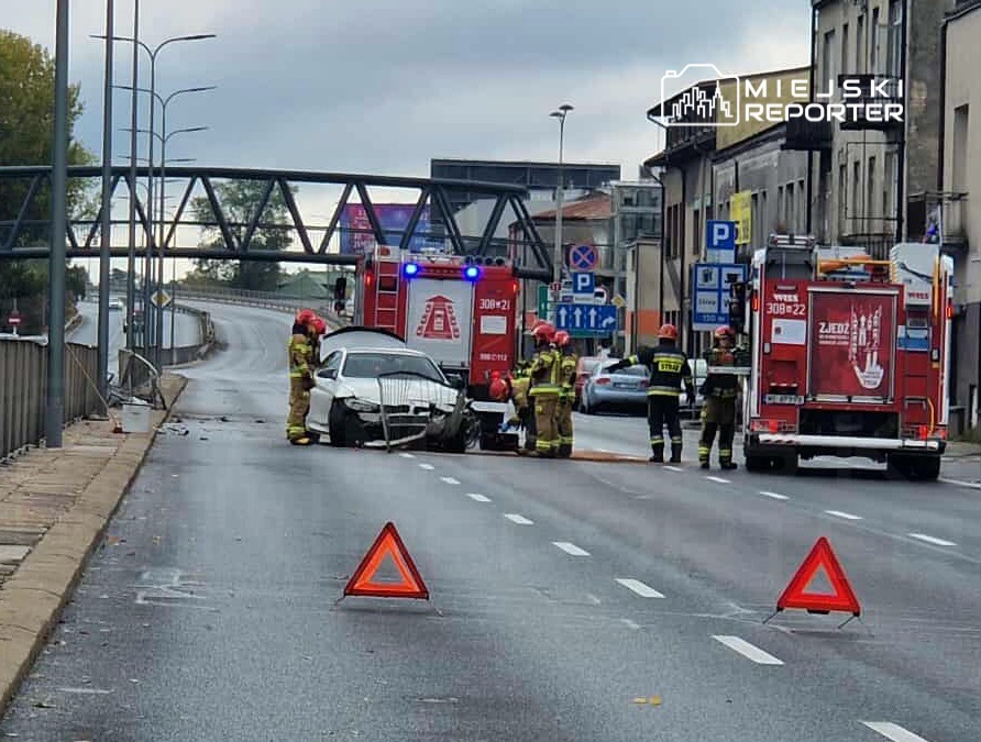 Grupa strażaków pracuje przy uszkodzonym samochodzie na jezdni, obok stoją dwa wozu strażackie.