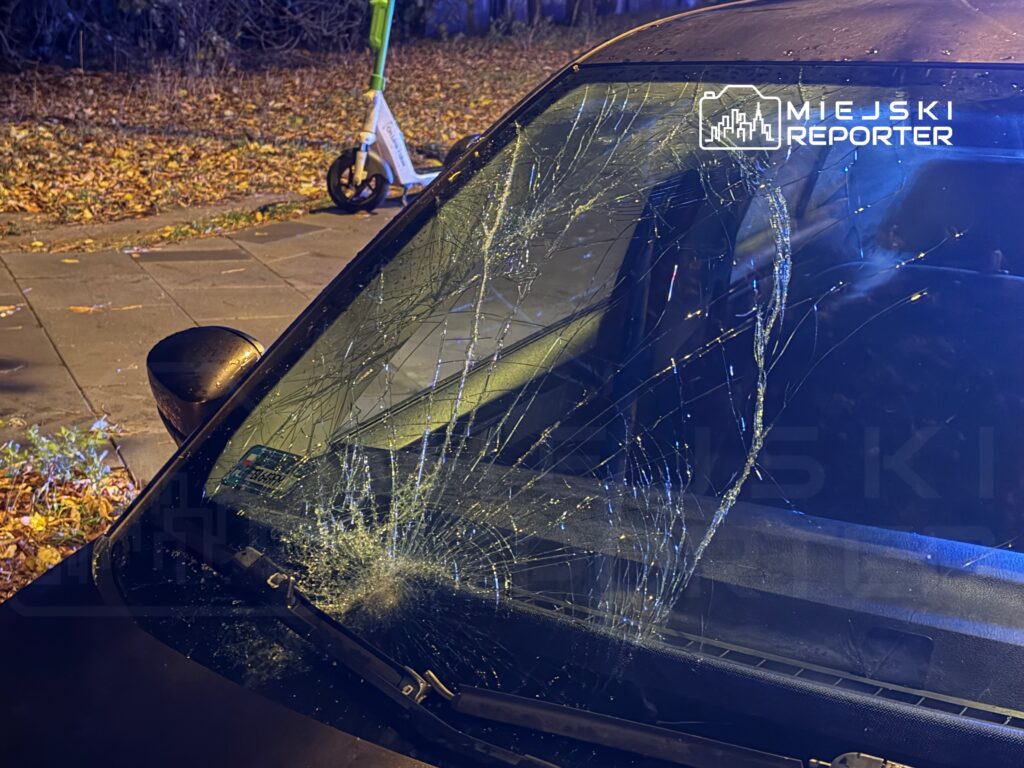 Pęknięta szyba przednia samochodu zaparkowanego na ulicy, w tle widoczna elektryczna hulajnoga i opadłe liście.