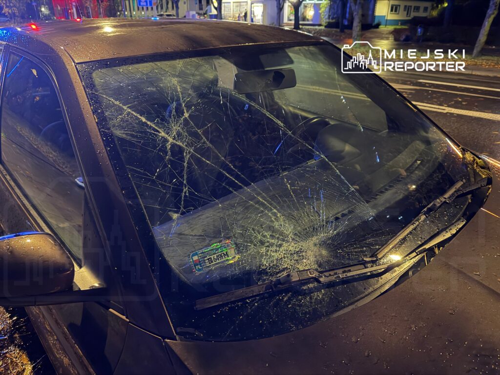 Uszkodzony samochód z pękniętą szybą przednią stojący na mokrej ulicy w nocy.