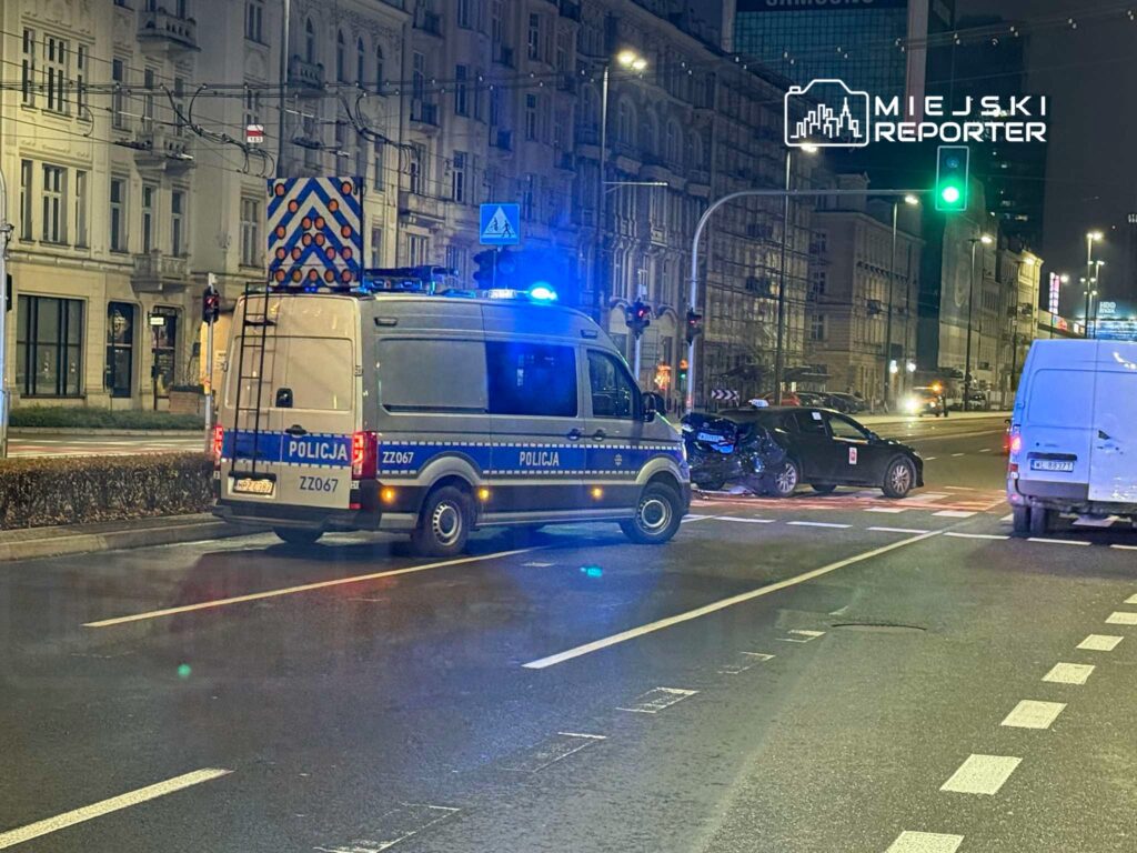 Policyjny van z włączonymi sygnałami świetlnymi stoi obok rozbitego samochodu osobowego na ulicy w nocy.
