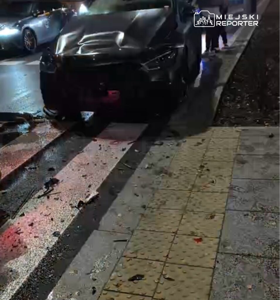 Czarny samochód osobowy z uszkodzoną maską stoi na pasach, obok widać rozbite szkło i resztki pojazdu.