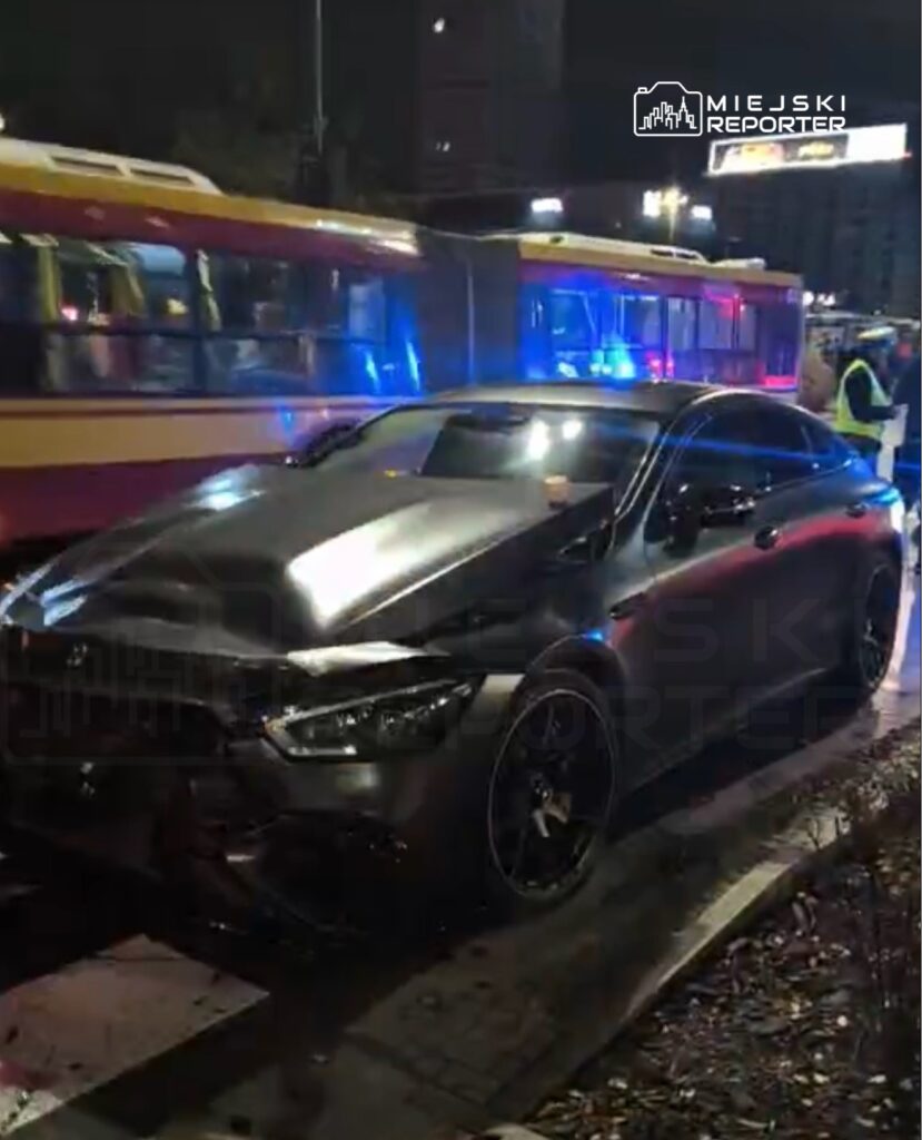 Czarny samochód osobowy z uszkodzoną przednią częścią stoi na poboczu obok tramwaju, w tle widać niebieskie światła policyjne.